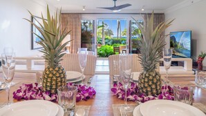 Une table à manger dressée avec des assiettes, des verres et deux ananas, donnant sur un jardin luxuriant et avec un téléviseur en arrière-plan.