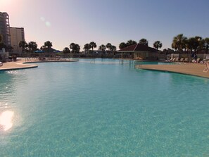 A large outdoor swimming pool with clear blue water, surrounded by palm trees and a building with a red roof.