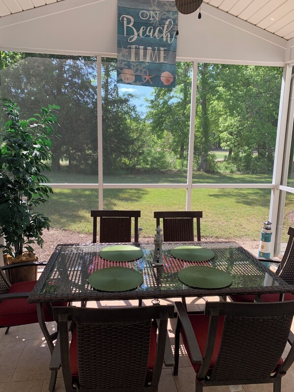 A covered patio with a table set for four, surrounded by wicker chairs, a hanging sign reading "ON Beach TIME", and a view of a grassy area with trees.