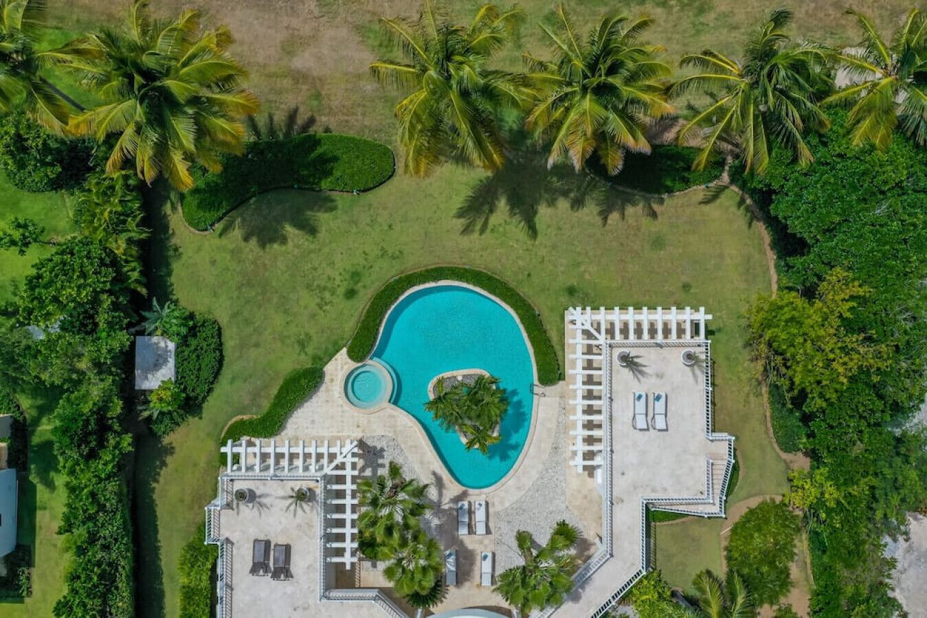 Vista aérea de un resort con una piscina grande, palmeras y un área cercada con sillas de descanso.