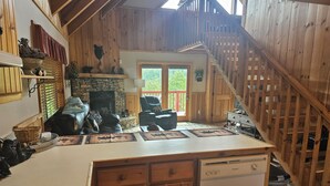Een rustiek blokhutinterieur met een stenen openhaard, houten wanden, een eethoek met tafel en stoelen, en een trap naar een hoger gelegen niveau.