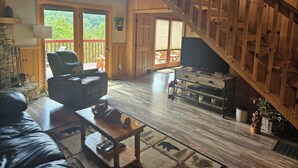 Een ruime woonkamer met een houten trap, een comfortabele bank, een televisie en een balkon met uitzicht op groen.