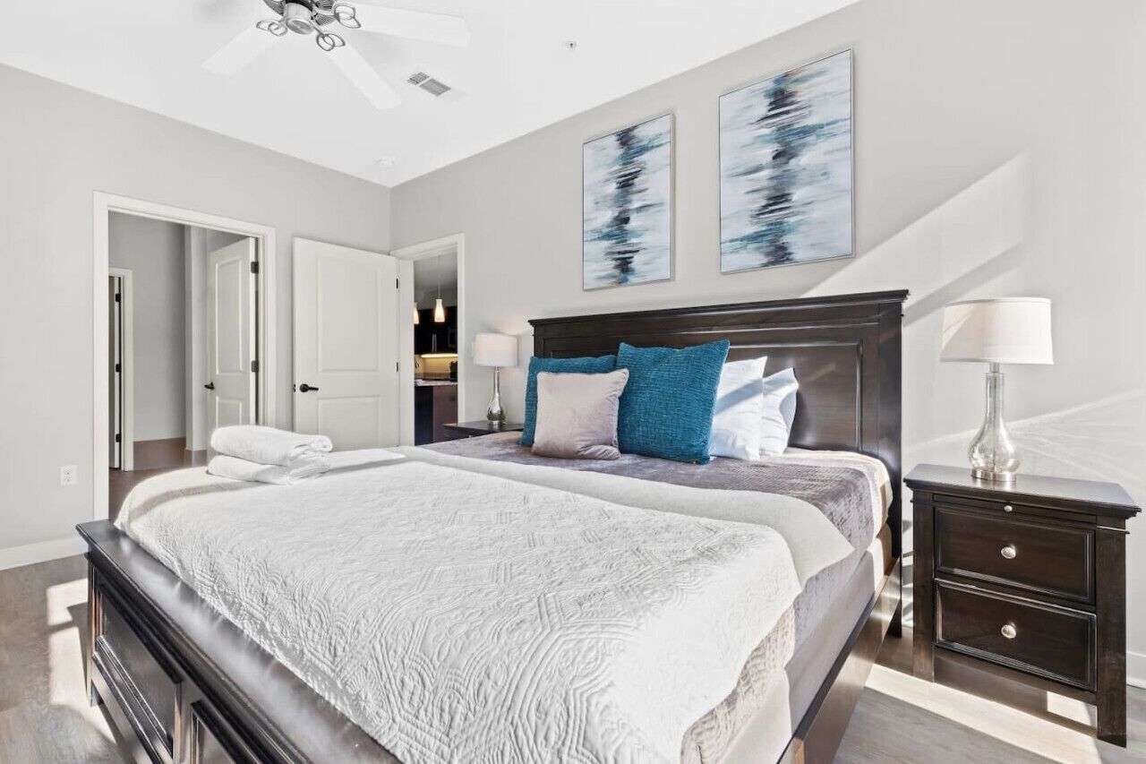 A bedroom with a large bed, a dark wooden headboard, and a nightstand with a lamp. There are two doors leading to other rooms.