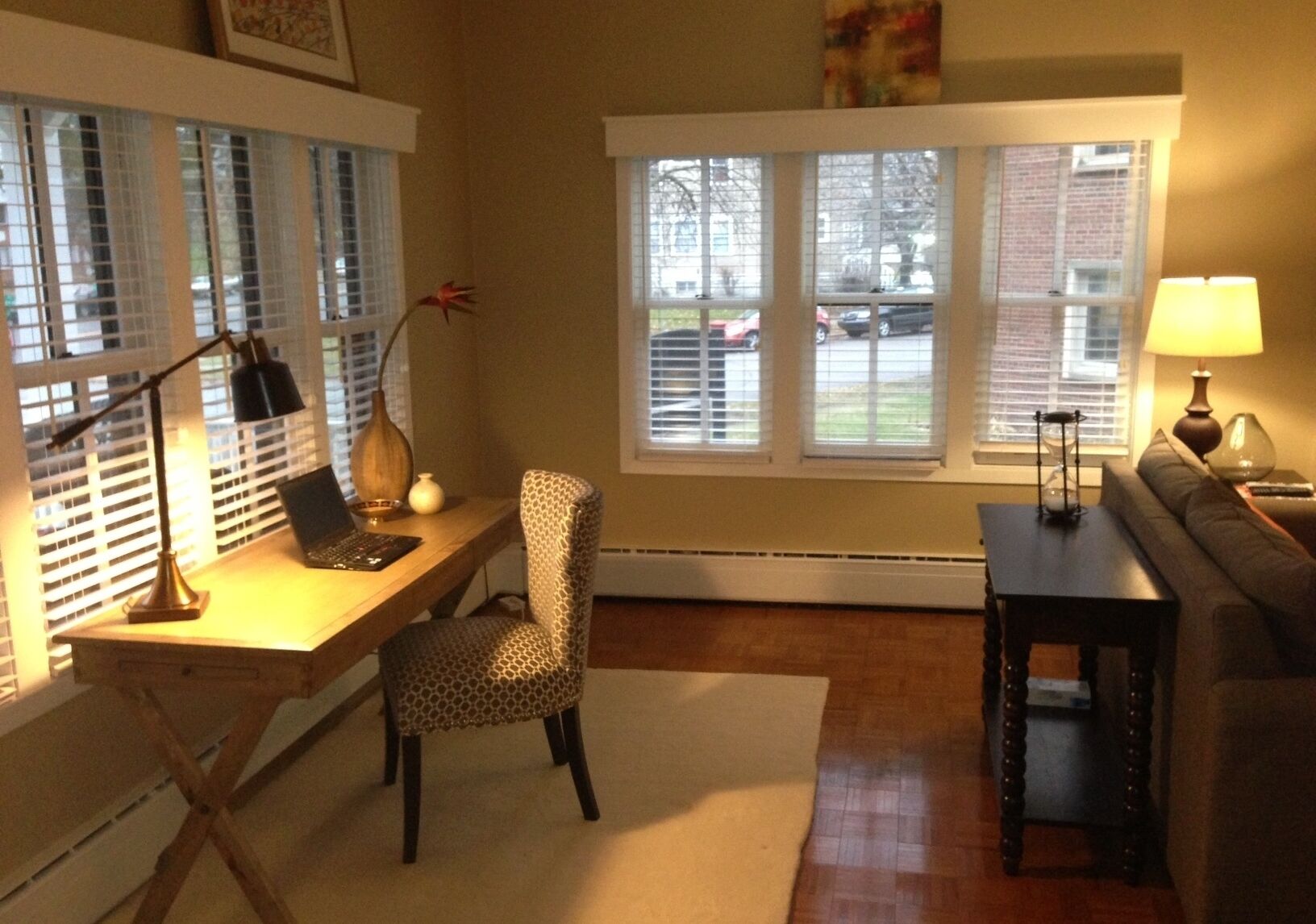 Ein gemütliches Wohnzimmer mit einem Holztisch, einem Sessel mit Leopardenmuster, einer Couch und einem Couchtisch. Zwei Fenster mit Jalousien, eine Lampe auf dem Tisch und eine Vase mit einer Pflanze sind ebenfalls vorhanden.