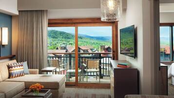 A two-bedroom villa with a living area featuring a sofa, coffee table, and artwork. There is a balcony with a view of the surrounding landscape.