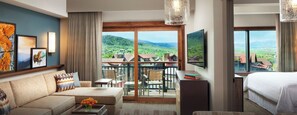 A two-bedroom villa with a living area featuring a sofa, coffee table, and artwork. There is a balcony with a view of the surrounding landscape.