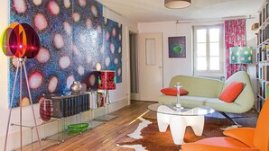 A living room with a glass coffee table, a sofa with orange cushions, a bookshelf, and a wall with a galaxy-themed artwork.