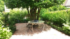 A garden patio with a round table and chairs, surrounded by lush greenery and a potted plant.