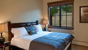 A neatly made bed with a dark headboard, two bedside lamps, and a window with a view of a park.