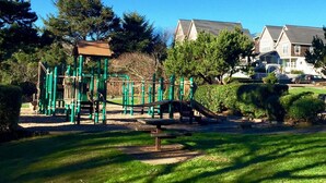 A playground with climbing structures and a slide, surrounded by grass and trees, with houses in the background.