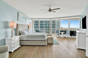 A bedroom with a bed, bedside tables, a ceiling fan, a large window with a view, and a balcony with seating.