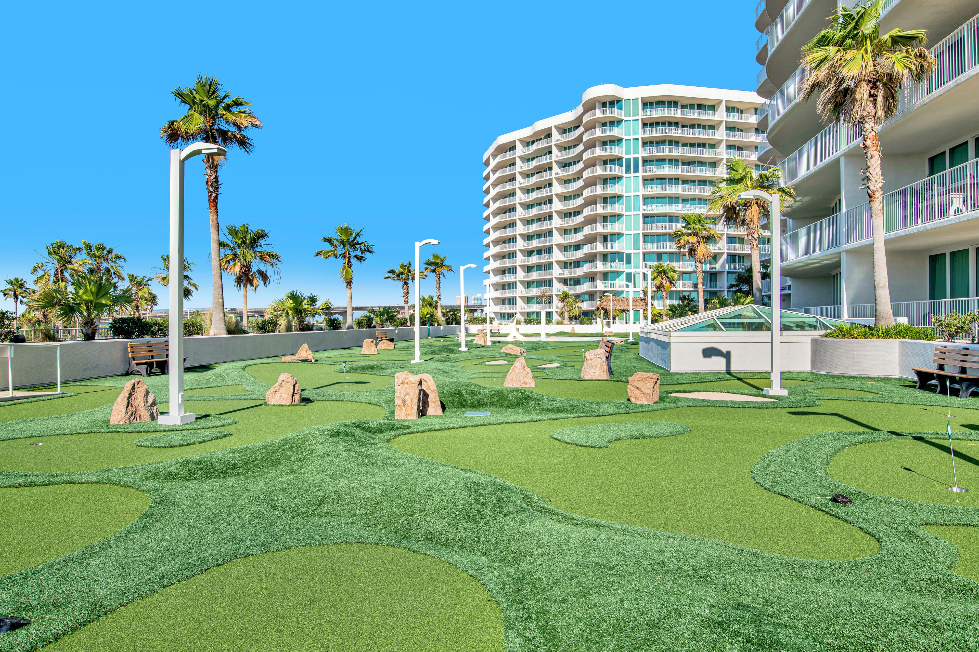 A hotel with a modern design, featuring a large building with multiple balconies and a well-maintained artificial green lawn with palm trees and rocks.