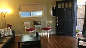A living room with a black coffee table, two white chairs with red cushions, a small white table with a framed welcome sign, a black door, and a window with blinds.