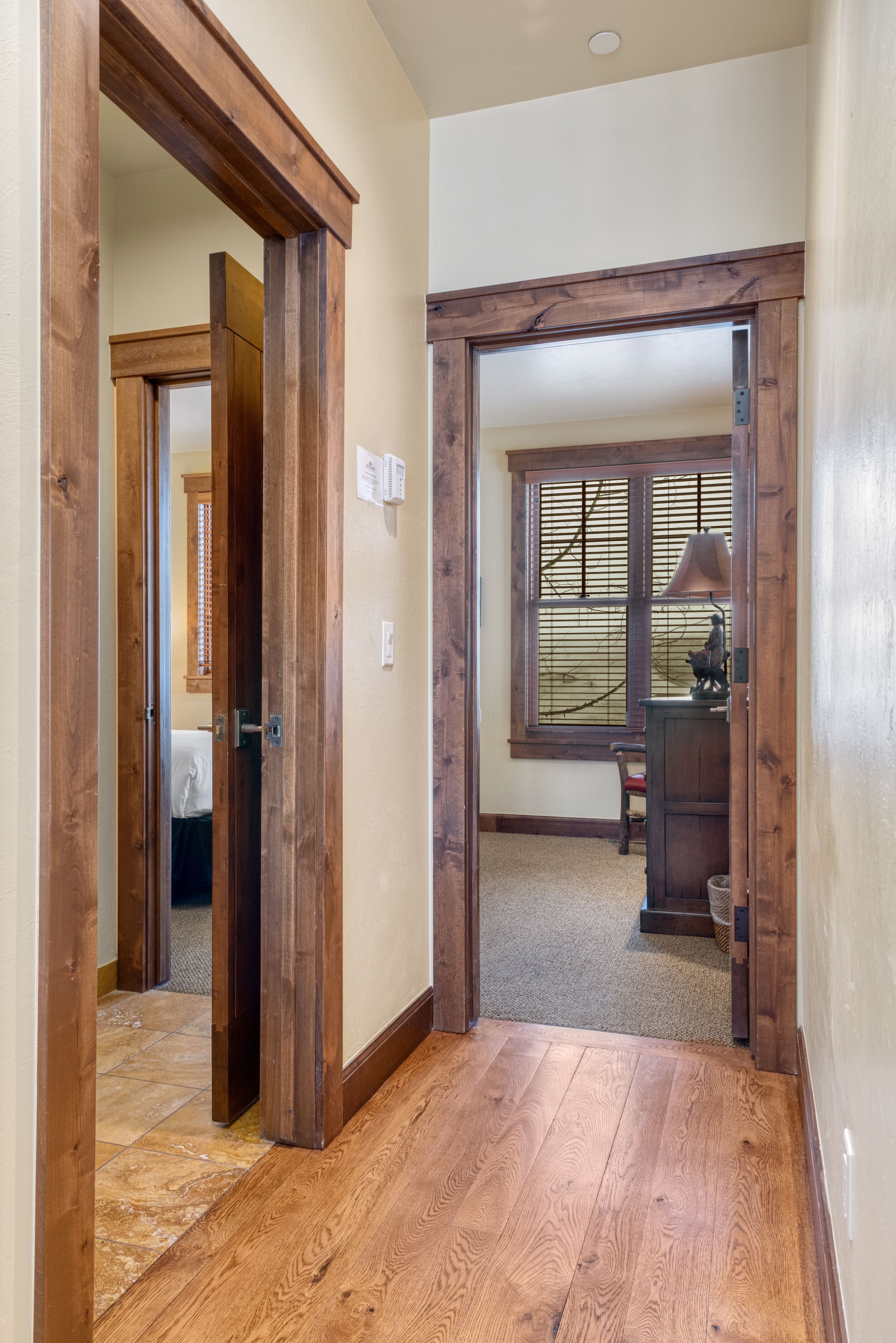 A hallway with wooden flooring and a door leading to a room with a window and a desk.