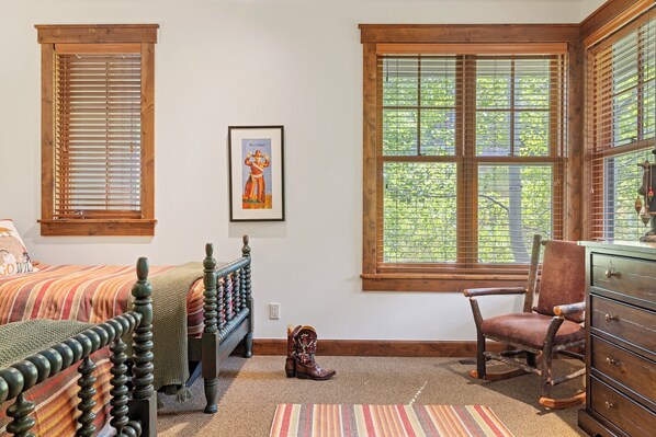 A bedroom with two beds, a rocking chair, a dresser, and large windows with blinds.