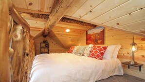 Un dormitorio con techo inclinado, cama con ropa de cama blanca, almohadas coloridas, mesita de noche con lámpara y un cuadro en la pared.