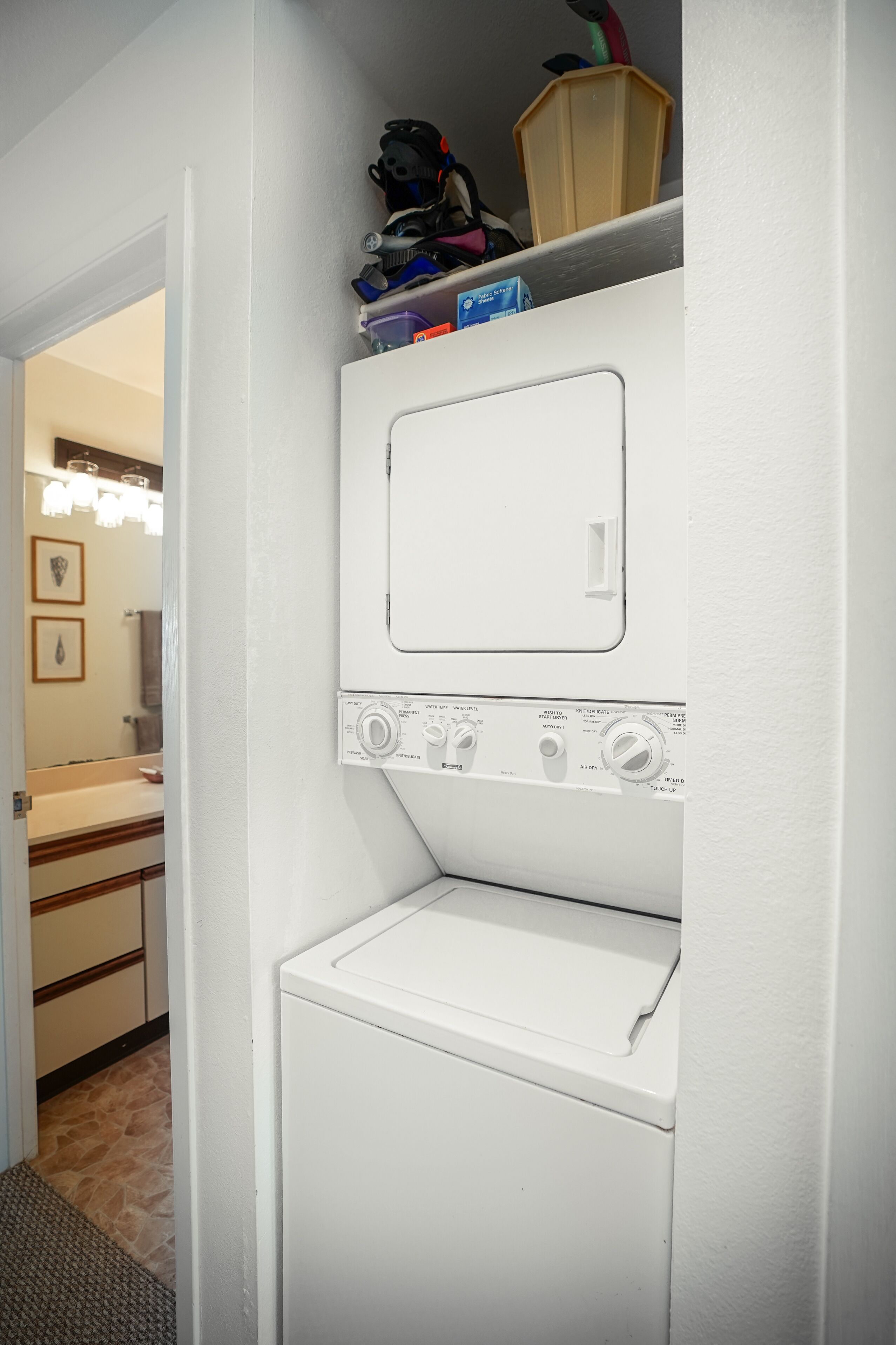 Une buanderie équipée d’un lave-linge et d’un sèche-linge à chargement frontal, d’une étagère au-dessus du sèche-linge où sont rangés divers objets, et d’une porte donnant accès à une salle de bain.