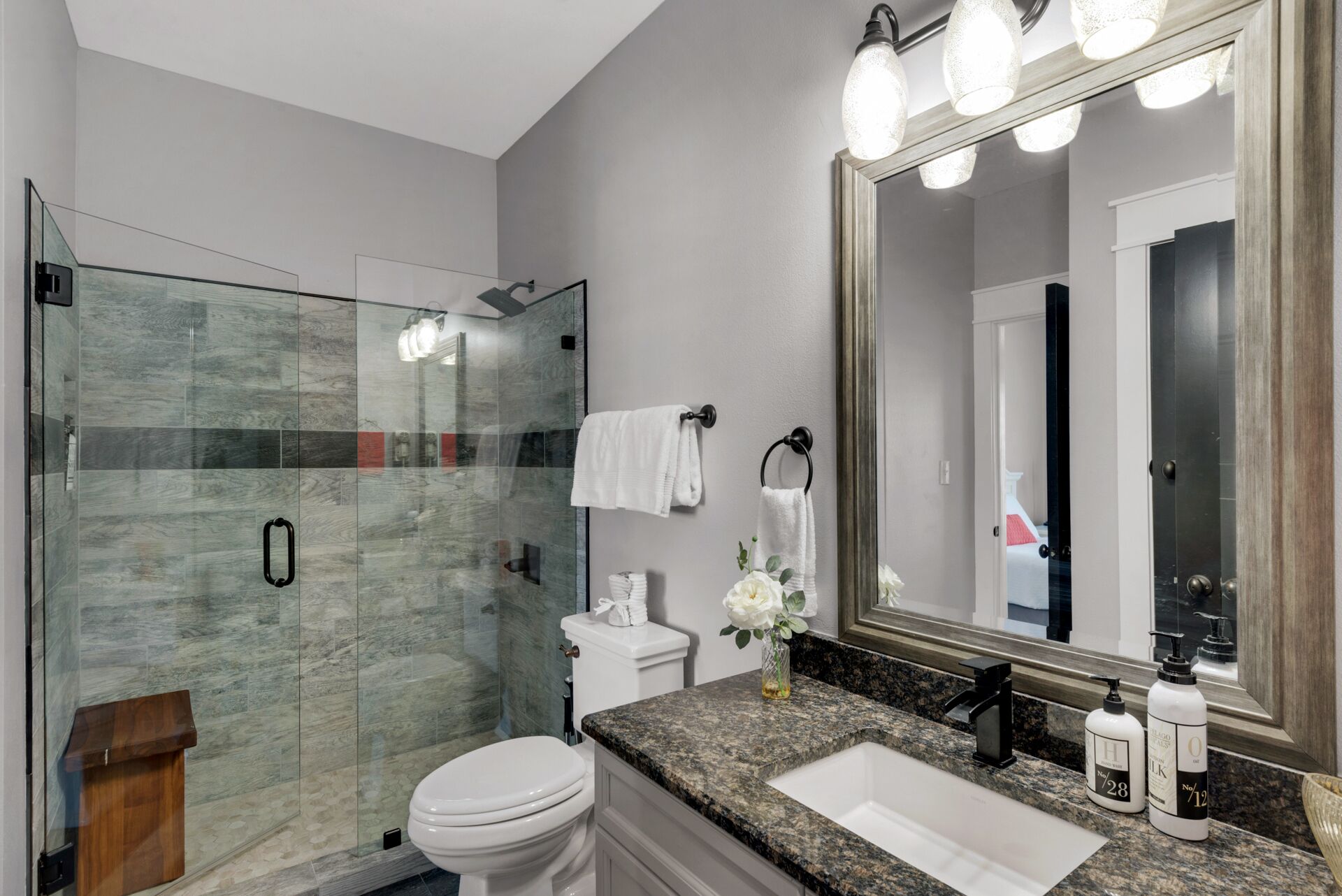 A modern bathroom with a glass shower enclosure, a white toilet, a granite countertop, and a large mirror with lights.