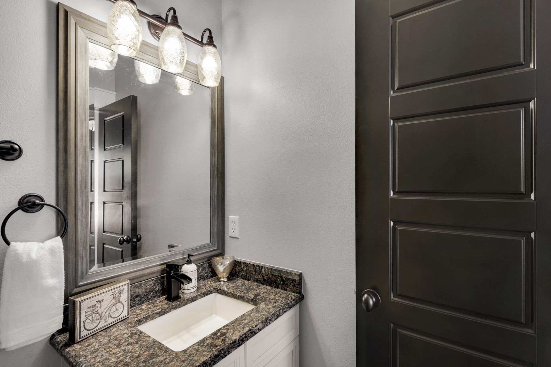 A bathroom with a granite countertop, a white sink, a mirror with lights, and a dark door.