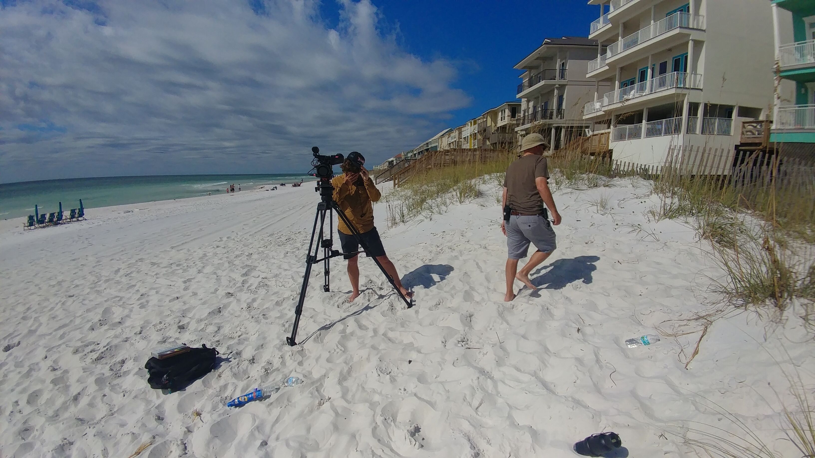 A beach with a camera on a tripod, a person walking, and buildings in the background.
