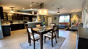 A modern kitchen with dark cabinets, a central island, and a dining area with a glass table and chairs. - Modern 3 bedroom, 2 bath getaway with HEATED private pool and jacuzzi (Fort Myers)