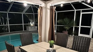 An outdoor dining area with a pool view, featuring a table and chairs, and large windows. - Modern 3 bedroom, 2 bath getaway with HEATED private pool and jacuzzi (Fort Myers)