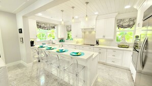 A modern kitchen with white cabinetry, a central island with bar stools, and large windows with views of greenery.
