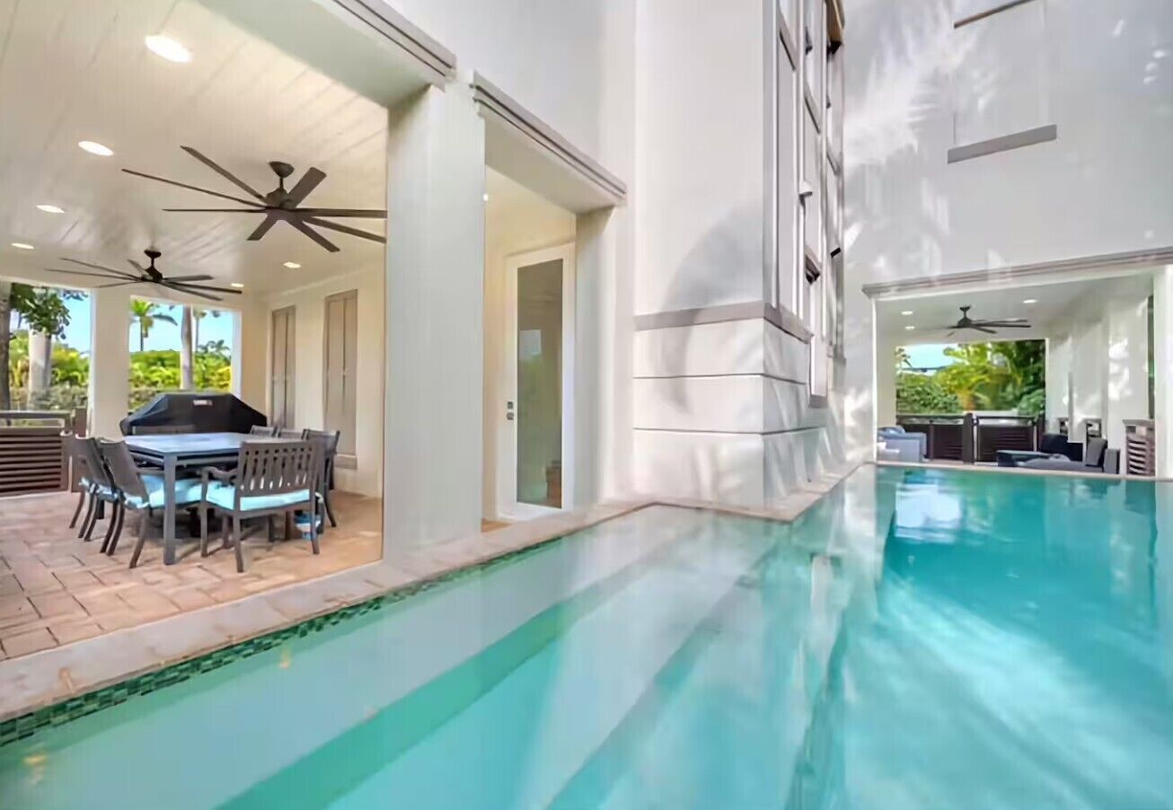 An indoor swimming pool with ceiling fans, a dining area with a table and chairs, and a view of the outdoors.