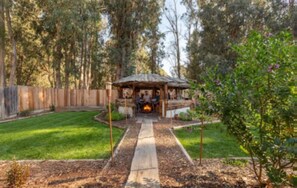 Un belvédère en bois avec un foyer au centre, entouré d’une pelouse bien entretenue et d’une clôture en bois.