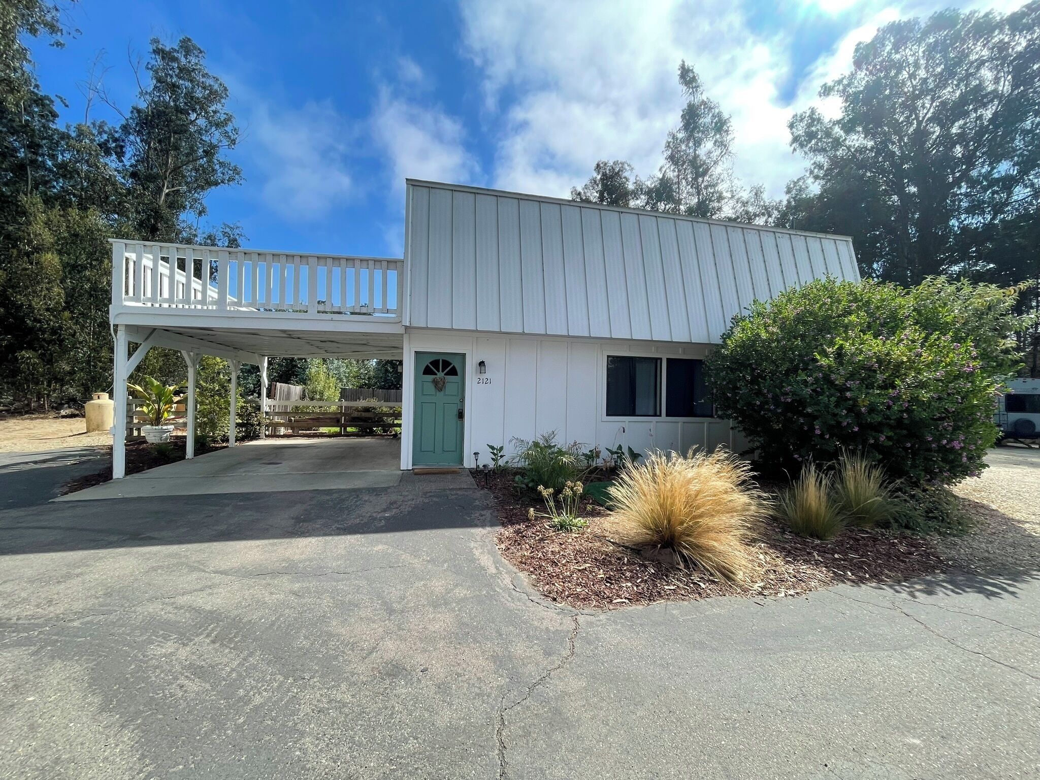 A two-story building with a covered porch and a green door, surrounded by landscaped garden beds and trees.