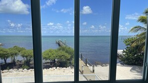 A view through a window of a beach with a wooden pier extending into the water, surrounded by greenery and a clear sky. - Sunset Catch- Gulf Front, Open Water Views, Private Pool and Dock, Kayaks. (Marathon)