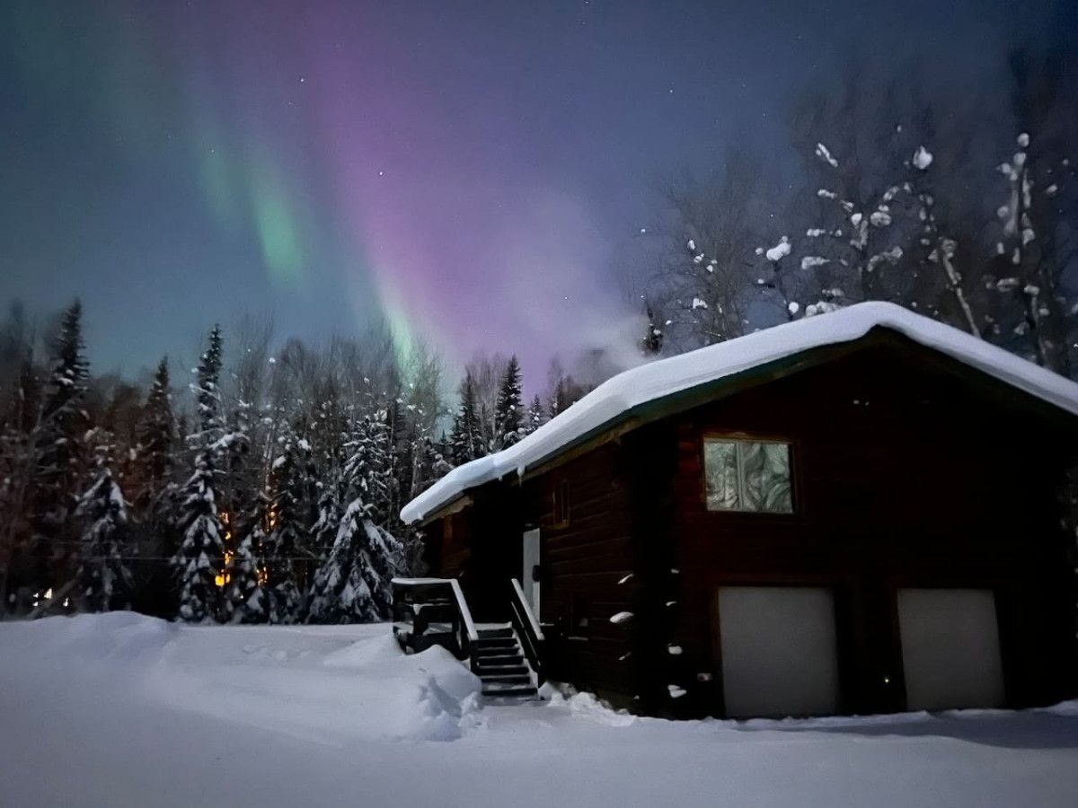 A cabin with a snow-covered roof, surrounded by snow-covered trees, under a night sky with aurora borealis.