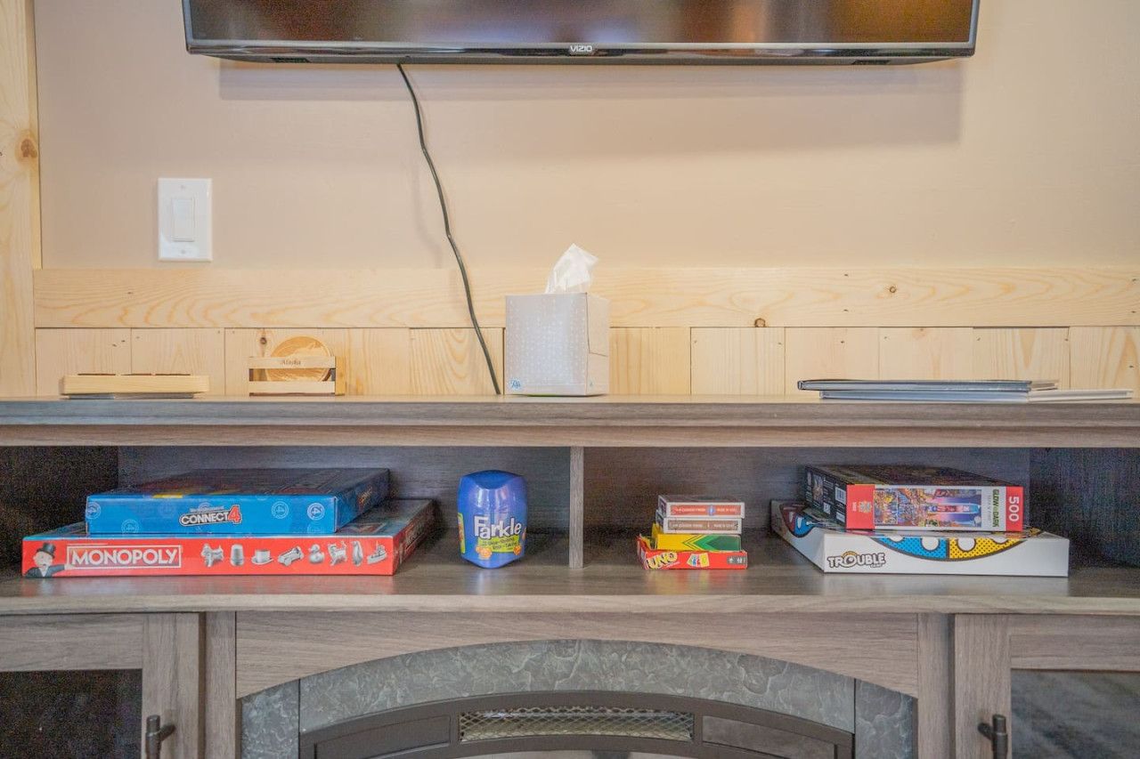 A room with a television mounted on the wall, a wooden shelf with various board games, and a fireplace below.