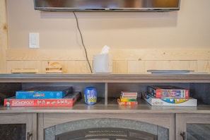 A room with a television mounted on the wall, a wooden shelf with various board games, and a fireplace below. - Tucked Away Timber Lower Suite (Fairbanks)