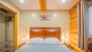 A bedroom with a wooden headboard, a bed with a blue quilt, bedside lamps, and a window with curtains. - Tucked Away Timber Lower Suite (Fairbanks)