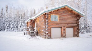 A log cabin with a snow-covered roof, a front door, and a small porch with a wooden bench. - Tucked Away Timber Lower Suite (Fairbanks)