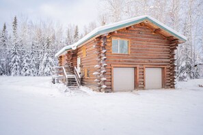 Une cabane en rondins dont le toit est recouvert de neige, avec une porte d’entrée et un petit porche agrémenté d’un banc en bois.