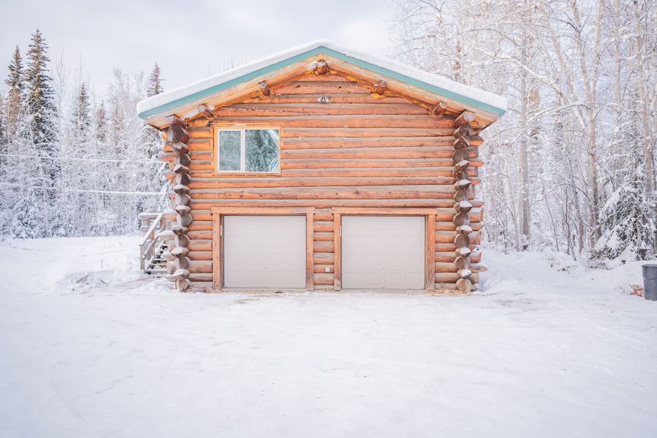Una cabaña de troncos con dos puertas de garaje, rodeada por un bosque nevado.