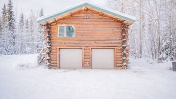 Una cabaña de troncos con dos puertas de garaje, rodeada por un bosque nevado.