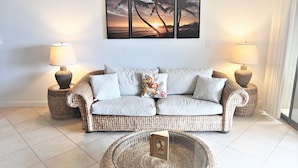 Uma sala de estar com um sofá trançado, duas mesinhas de cabeceira com luminárias, uma mesa de centro de vidro e um tríptico de pintura fixado na parede.