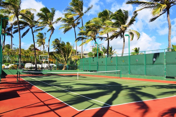 Un court de tennis à la surface verte et rouge, entouré de palmiers et d’une clôture verte.