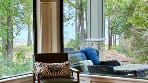 Ein Zimmer mit einem großen Fenster, einem Holzstuhl mit gemustertem Kissen, einem Korbssessel mit blauen Kissen und einem Steinkamin.
