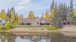 A two-story house with a front deck, a small pond in front, and a forested area in the background.