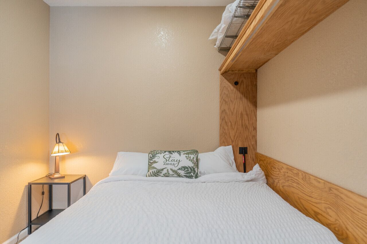 Ein ordentlich gemachtes Bett mit weißer Decke und einem Kopfkissen mit dem Aufdruck 'Stay Awake', einer Nachttischlampe und einem Holzkopfteil.