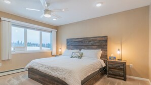 A bedroom with a large bed, a ceiling fan, bedside lamps, and a window with a view of trees.