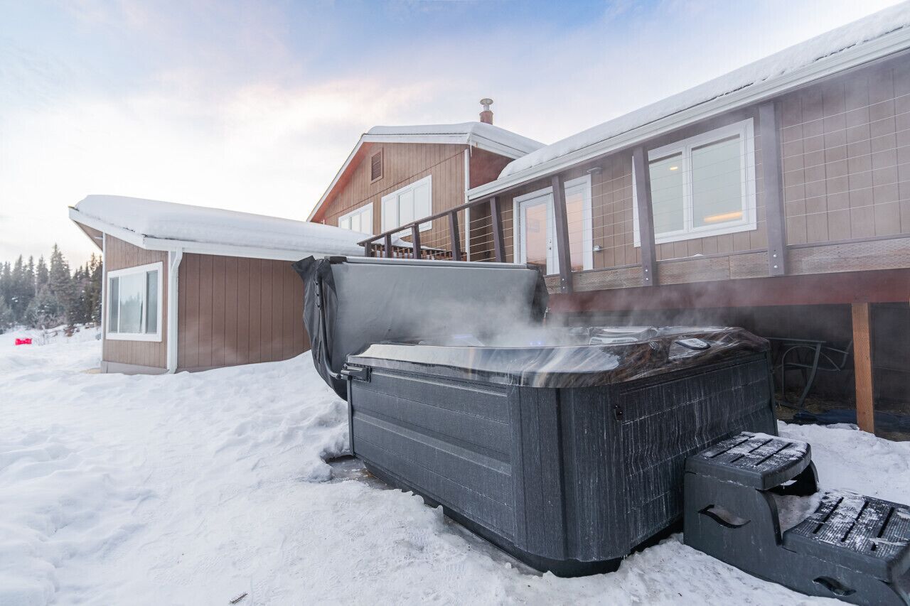 A hot tub in a snowy outdoor area with a wooden house in the background.