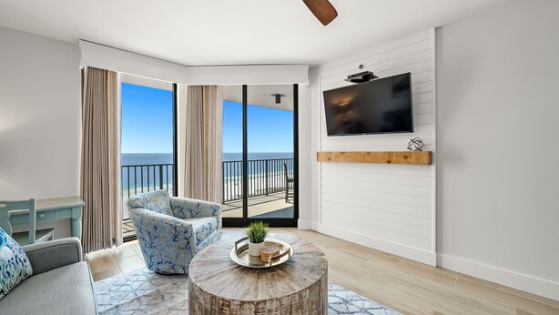 A modern living room with a large window, a ceiling fan, a flat-screen TV, and a wooden coffee table. - Escape to the Beach! Phoenix 8, 1402, 2 BR/2 Bath (Orange Beach)