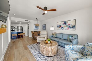 A modern living room with a blue sofa, a round coffee table, and a dining area with a chandelier. - Escape to the Beach! Phoenix 8, 1402, 2 BR/2 Bath (Orange Beach)
