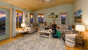 Ein gemütliches Wohnzimmer mit Deckenventilator aus Holz, einer Sofagarnitur, einem Couchtisch und Blick auf Berge durch die Fenster.