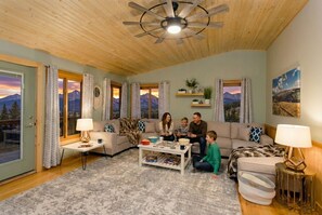 Una sala de estar acogedora con un ventilador de techo de madera, un conjunto de sofás, una mesa de centro y vistas a las montañas a través de las ventanas.